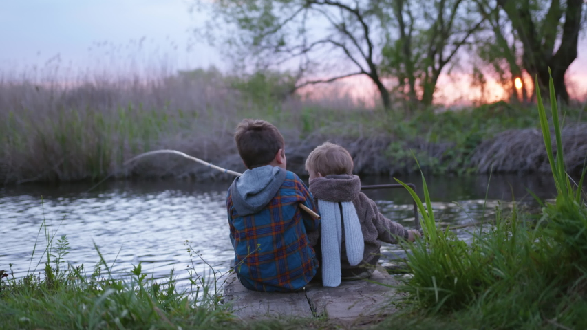Family weekend, fishermen little brothers sit on pier and fish with a wooden fishing rod in stream in middle on sunset on fun holidays outside city among reeds, back view | Shutterstock HD Video #1052841941