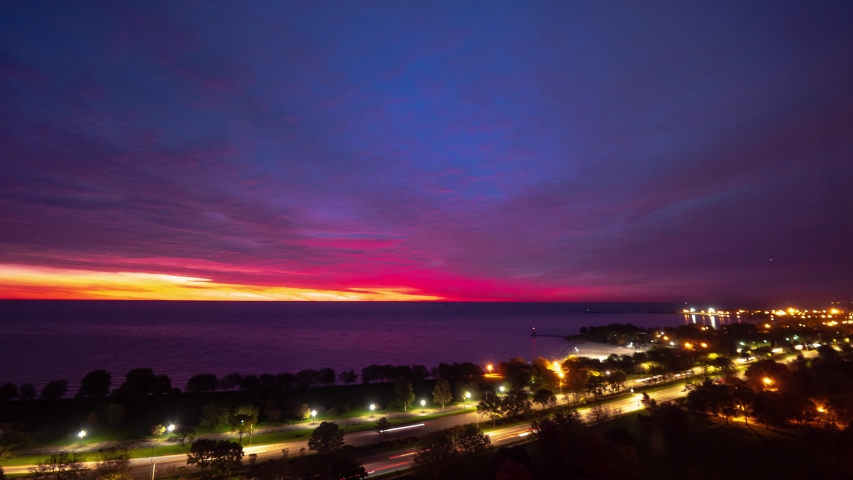 A beautiful zoom out aerial sunrise time lapse with sky on fire pink, yellow, blue, purple, orange and red eruption of color on the clouds as traffic builds on Lake Shore Drive below in Chicago.