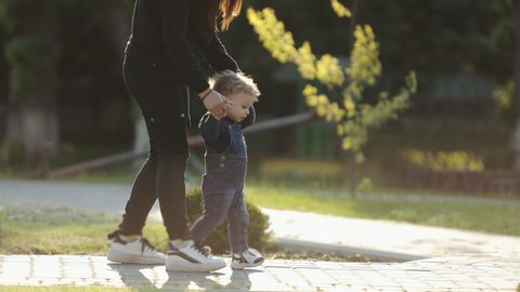 Cute child walking with mother in the park, mom holding hand baby boy. Mother supports child making first steps on park holding hand.