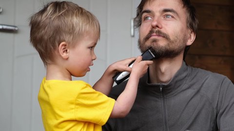 Little child boy cut father beard with clipper. Family haircut, hygiene, beauty and selfcare at home during quarantine lockdown, lifestyle concept.