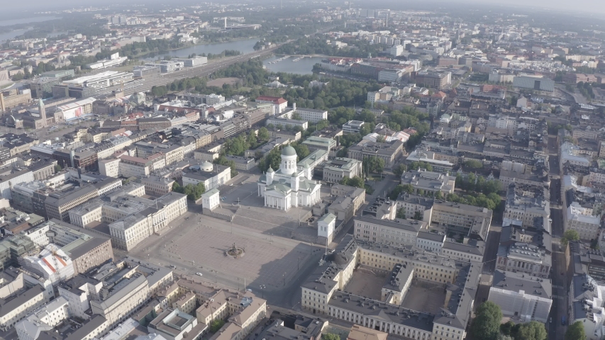 Helsinki, Finland. The Helsinki Cathedral dominates the neoclassical Senate Square from an elevated aerial perspective, its white facade and green copper dome standing in sharp contrast against the surrounding urban grid. The camera executes a smooth, sweeping arc around the cathedral, gradually revealing the symmetrical Empire-style ensemble designed by Carl Ludvig Engel, including the Government Palace and University of Helsinki main building that frame the square. The footage captures the cathedral