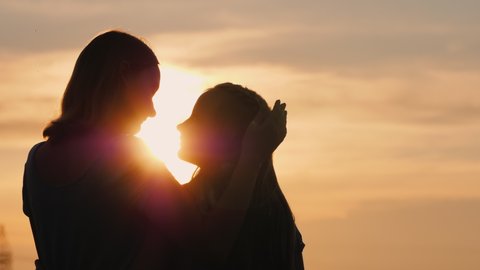 A woman gently kisses her daughter, silhouettes against the sky where the sun sets