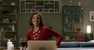 Beautiful woman in a red dress having a glass of wine chatting, online date with partner. Stay home, quarantine life. Shot with anamorphic lens