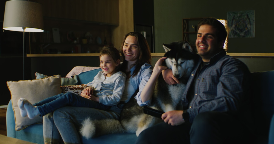 Family of three - father, mother and daughter wathcing TV in the evening. Pet husky dog lying near them. Togetherness, quarantine, stay home Royalty-Free Stock Footage #1052858204
