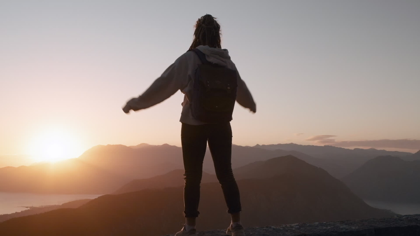 Young inspired Dreadlocks woman raises her hands up standing on the top of a mountain above the sea against beautiful sunrise sky. Watching the sunset with beautiful landscape in Montenegro.
 | Shutterstock HD Video #1052859839