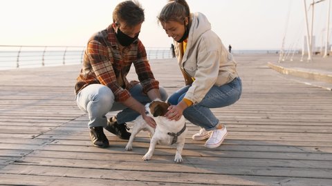 Young happy couple in protective medical masks petting cute Jack Russel terrier dog outdoors near the sea, slow motion