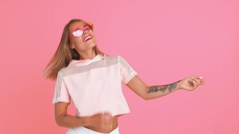 Young lady with shiny freckles and tattooed hands, in sunglasses and top. Acting like playing guitar, laughing, posing on pink background. Close up