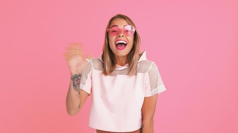 Woman with shiny freckles, in sunglasses and top. Waving hands, saying hi, smiling, sending you an air kiss, posing on pink background. Close up
