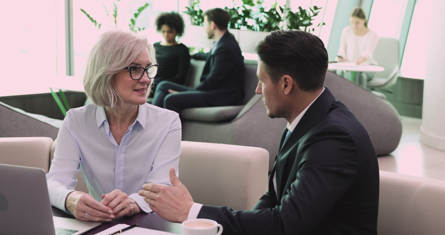 Friendly different generation business people discussing partnership conditions or project in modern coworking space. Confident middle aged female leader talking to male employee in formal wear.