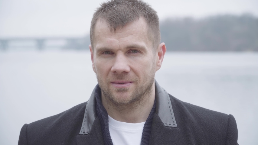 Close-up of serious brunette Caucasian man with grey eyes posing outdoors. Portrait of confident young guy in elegant coat standing on riverbank on cloudy day. Lifestyle, confidence, leisure.