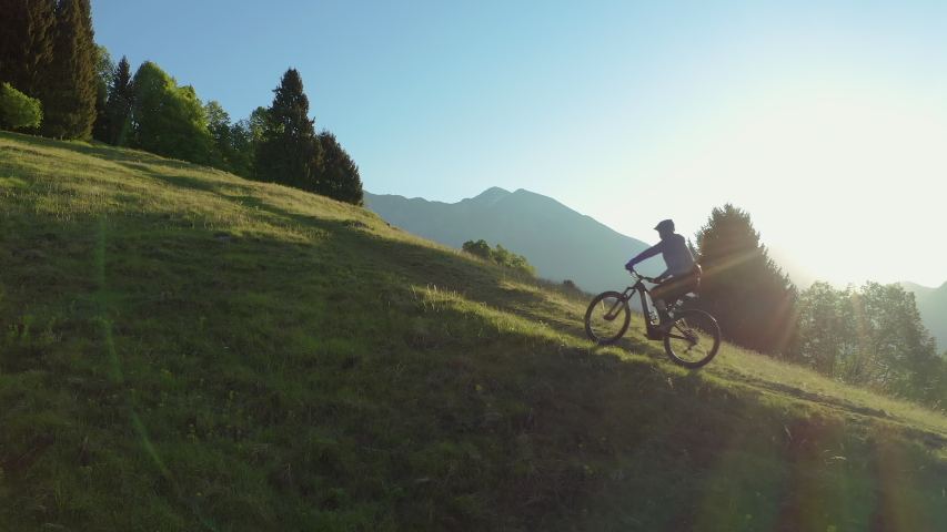 Drone is flying along an athletic man pedalling an MTB E-bike up a steep grassy hill. Beautiful view of the mountains at sunrise/sunset with sun flare. Alone in nature, thinking about life. | Shutterstock HD Video #1052869115