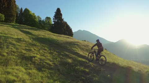 Drone is flying along an athletic man pedalling an MTB E-bike up a steep grassy hill. Beautiful view of the mountains at sunrise/sunset with sun flare. Alone in nature, thinking about life.