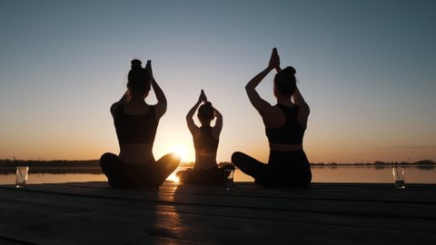Silhouette of Diverse Group of Three Female Doing Yoga Awe Exercise Sitting in Lotus Position Together on Boardwalk Near Calm Water at Dusks Outdoor. Tranquil Namaste for Emotional Relief, Stress Free