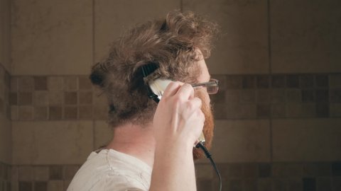 Man using electric clippers to cut his own hair at home during lock down.  Adult white male with beard and glasses in the middle of cutting his own hair with trimmer, while barbers are closed.
