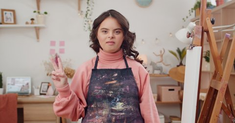 Adorable teen with down syndrome wearing in artists apron showing peace sign while looking to camera. Portrait of cute talanted girl posing while sitting near molbert in her room