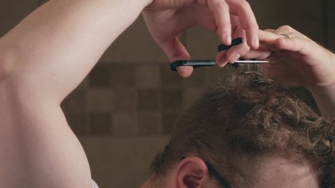 Close up of adult man using scissors to trim long portions of his own hair during the corona virus stay at home order.  White male DIY hair cut in home bathroom.