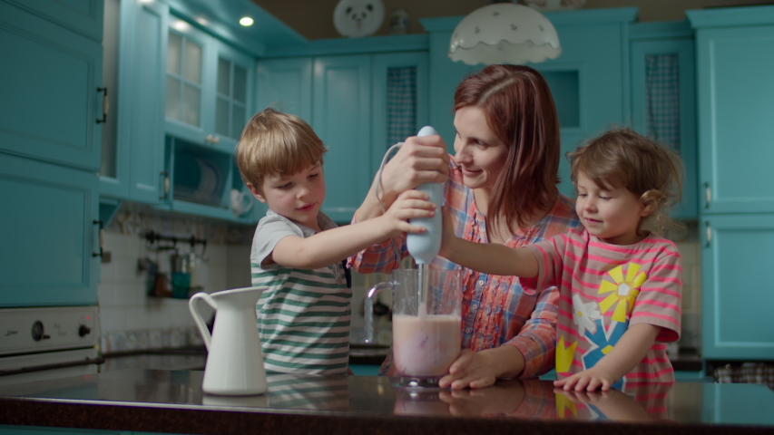 Happy family of mother and two kids cooking smoothie of strawberries, banana, vegan milk and yogurt with blender at home blue kitchen. Boy and girl helping mom to blender healthy cocktail.  Royalty-Free Stock Footage #1052887034