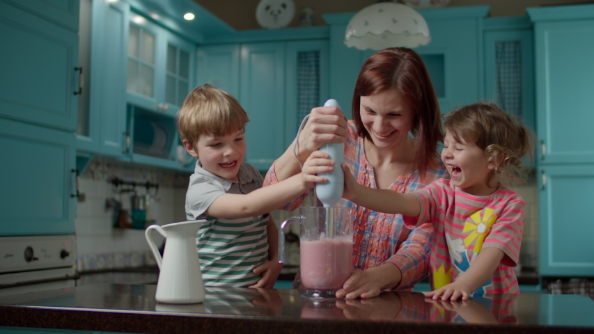 Happy family of mother and two kids cooking smoothie of strawberries, banana, vegan milk and yogurt with blender at home blue kitchen. Boy and girl helping mom to blender healthy cocktail.  Royalty-Free Stock Footage #1052887034