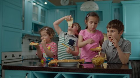 Group of four mixed age kids eating homemade pizza and potatoes standing on blue kitchen at home. Two boys and two girls enjoy eating pizza with hands.