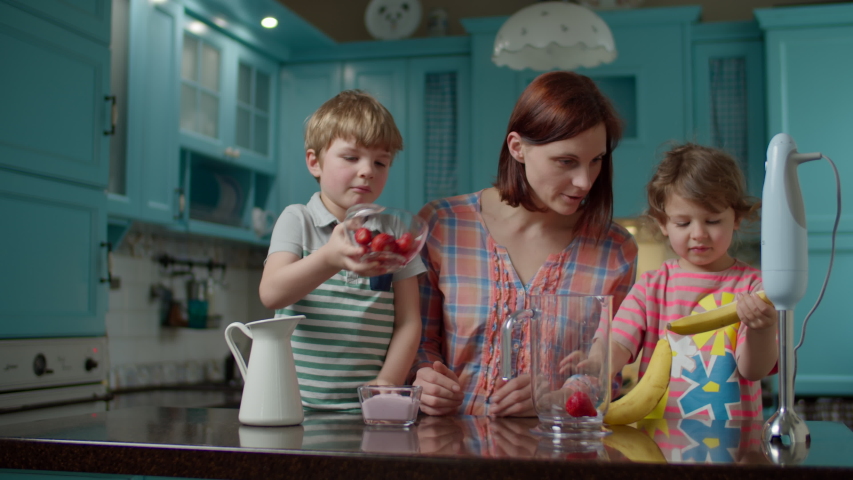 Happy family of mother and two kids cooking smoothie of strawberries, banana, vegan milk and yogurt with blender at home blue kitchen. Boy pouring berries and yogurt in bowl, girl peeling banana. Royalty-Free Stock Footage #1052887061