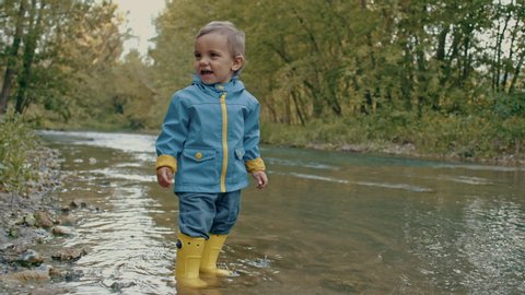 Cute baby in raincoat and rubber boots has fun by river, throws pebbles and laughs. Funny boy learns and explores natural world. Family, kids, love concept.