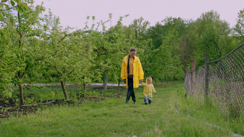 Mother and cute baby boy in yellow raincoat walking in green garden after rain. Child holds mom