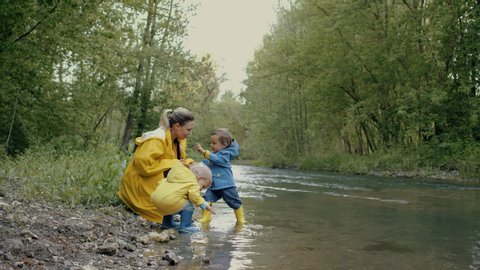 Mother and cute babies twins in raincoats and rubber boots has fun by river, throws pebbles and laughs. Funny boys learns and explores natural world. Family, kids, love concept.