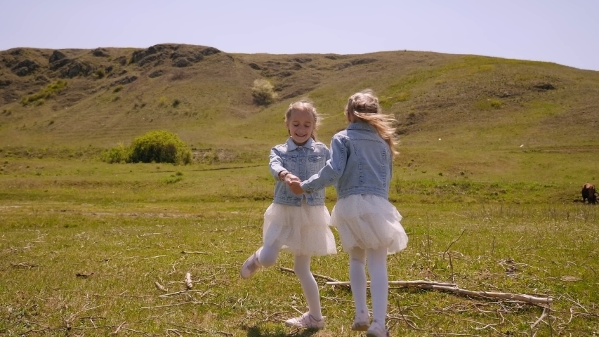 Two cheerful twin sisters, dressed in identical dresses, swirl holding hands on the field with green grass against the backdrop of the mountains. Slow motion.