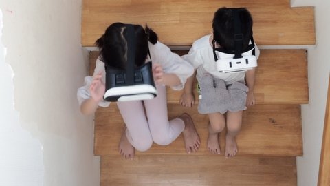 Sibling Asian school kids sitting at the stair at home while using VR glasses.Metaverse concept.