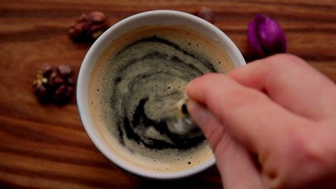 A man stirs sugar in a cup of aromatic coffee. A cup of aromatic coffee is placed on a wooden table. Man hands stir a coffee drink with a spoon.