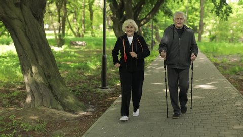 Active senior elderly couple 80 years old. Caucasian man training Nordic walking with ski trekking poles and woman is running. Park green tree background. Active family leisure. Healthy lifestyle