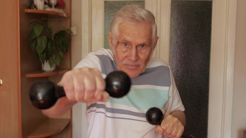 Senior elderly 80 years old caucasian man in sportswear stay in living room doing sports boxing exercises with dumbbells. Active old retiree exercising in the morning. People leisure healthy lifestyle
