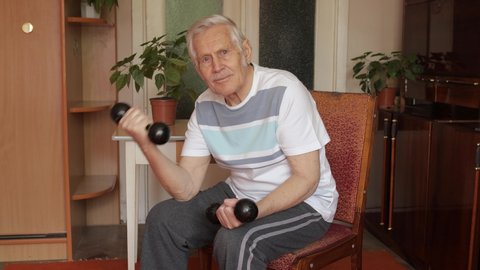 Senior elderly caucasian man in sportswear sitting on chair in living room, doing weight lifting dumbbell exercising at home. Active old retiree exercising in the morning. Leisure healthy lifestyle