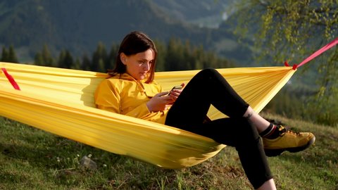 Relaxed Young Woman with Curly Hair Looking at Mobile Phone in Hammock and enjoys the scenery in the mountains