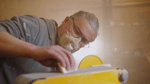 Masked carpenter use electric sander to polish wooden part of furniture. Wood craftsman working on woodworking machine.