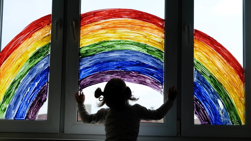 Little girl on background of painting rainbow on window. Kids leisure at home. Positive visual support during quarantine Pandemic Coronavirus Covid-19 at home.  Royalty-Free Stock Footage #1052895383