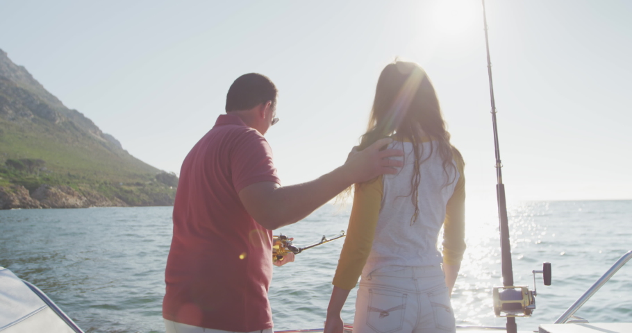 Rear view of a Caucasian man and his teenage daughter enjoying time together on holiday in the sun by the coast, embracing, holding and using fishing rods, in slow motion Royalty-Free Stock Footage #1052896157