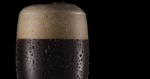 Glass of black beer on a black background. Beer sways in the glass, bubbles and foam rise. Glass of beer rotates slowly clockwise.