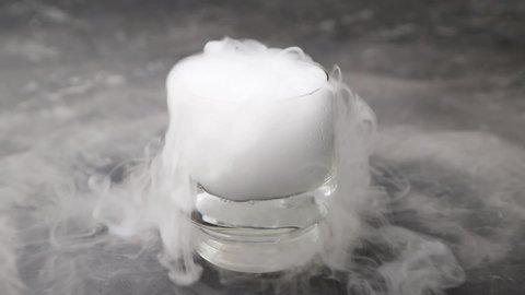 Dry ice fog flow from the transparent drinking glass on black concrete background. Chemical reaction with water. Side view, close up.