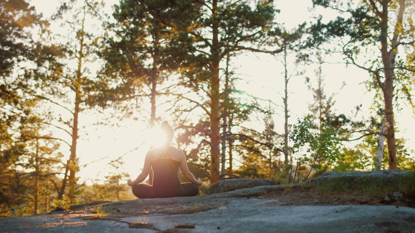 Woman practises yoga at sunrise in pine forest. Silhouette of young spiritual girl meditates in lotus pose nature outdoors, slow motion. Concept of fitness, healthy lifestyle, sun lens flare. | Shutterstock HD Video #1052897330