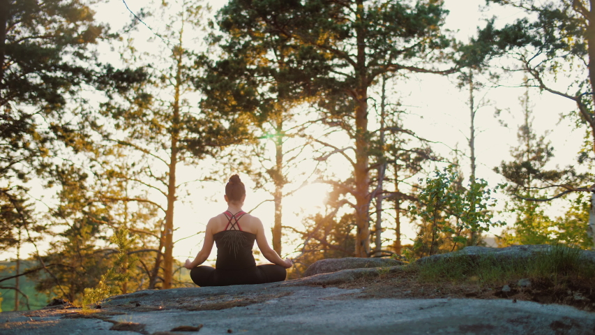 Woman practises yoga at sunrise in pine forest. Silhouette of young spiritual girl meditates in lotus pose nature outdoors, slow motion. Concept of fitness, healthy lifestyle, sun lens flare. Royalty-Free Stock Footage #1052897330