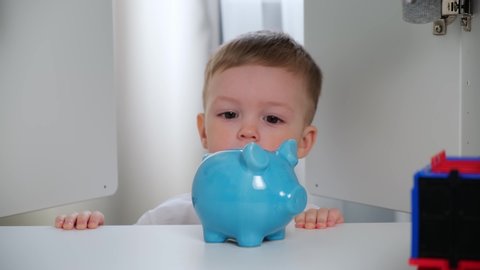 Small cute boy in white t-shirt tries to put green paper money in blue ceramic piggy Bank, money does not stick and bends. taken from first person. POV view from inside wardrobe. Money investment.