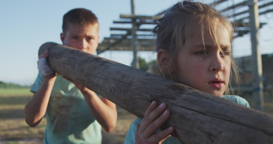 Front view of a happy Caucasian girl and boy on a boot camp on a sunny day, carrying a wooden post, in slow motion