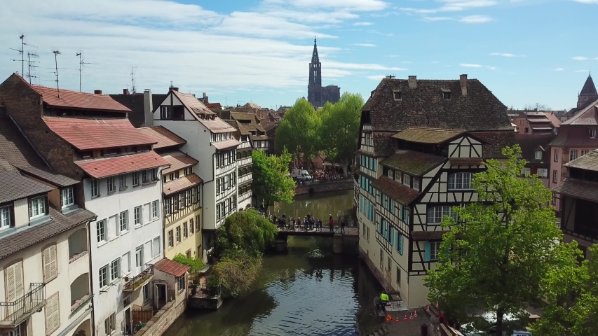 Drone flying over beautiful river and small bridge, surrounded by old houses and green trees in Petite France. Cathedral of Notre Dame on the background. Strasbourg, France | Shutterstock HD Video #1052902079