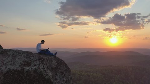 A young man works on a laptop outdoors on a mountaintop at sunset.