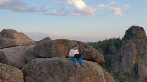 Drone footage of a young man working on a laptop sitting on a rock ledge in the mountains at sunset.
