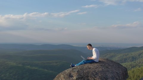 Drone shot of a young male freelancer sitting on top of a mountain and working on a laptop.