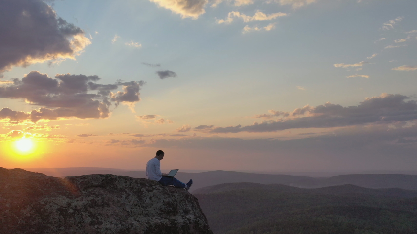 Aerial view of a young male freelancer working on a laptop sitting on a rock ledge in the mountains at sunset. Royalty-Free Stock Footage #1052907107