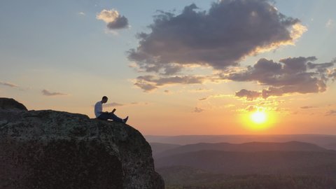 Aerial view of a young male freelancer working on a laptop sitting on a rock ledge in the mountains at sunset.