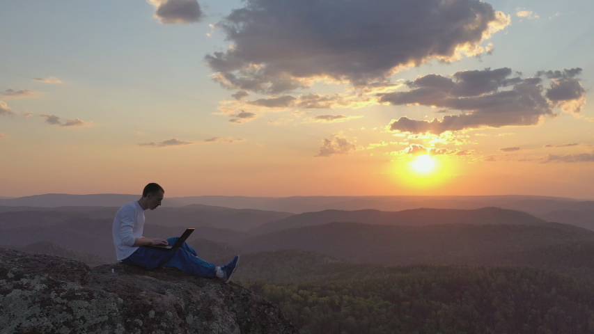 Epic drone shot of a silhouette of a young freelancer with a laptop on the edge of a rock ledge in the mountains. | Shutterstock HD Video #1052907110